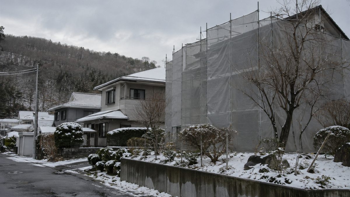 雪の残る住宅街で足場とシートに覆われた家